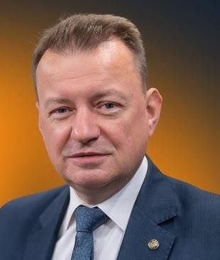 Mariusz Błaszczak podczas wystąpienia publicznego, z mikrofonami na pierwszym planie, symbolizującego jego aktywność polityczną.