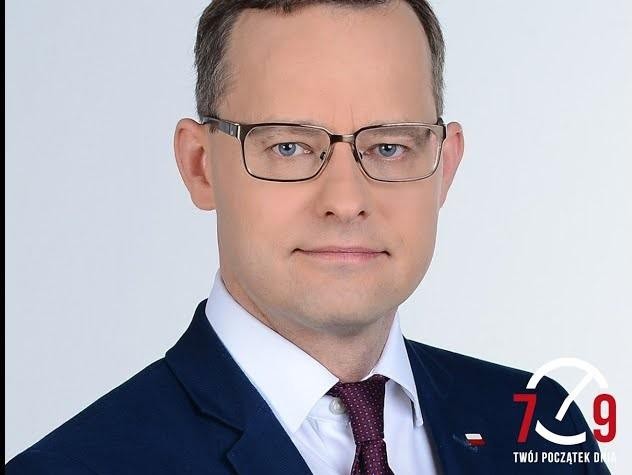 Zbliżenie na twarz Marcina Romanowskiego, z zdeterminowanym wyrazem, w tle subtelne elementy architektoniczne sugerujące instytucję publiczną.