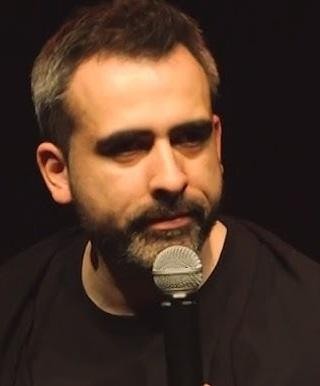 Abelard Giza na scenie podczas występu stand-upowego, dynamiczne ujęcie w trakcie mówienia, z mikrofonem w dłoni, w tle widownia.