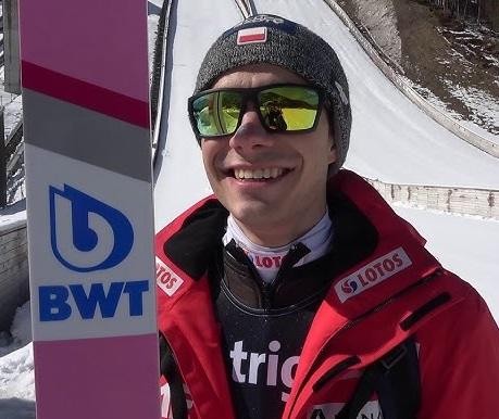 Jakub Wolny uśmiecha się, trzymając kask narciarski, na tle rozmytego krajobrazu treningowego.